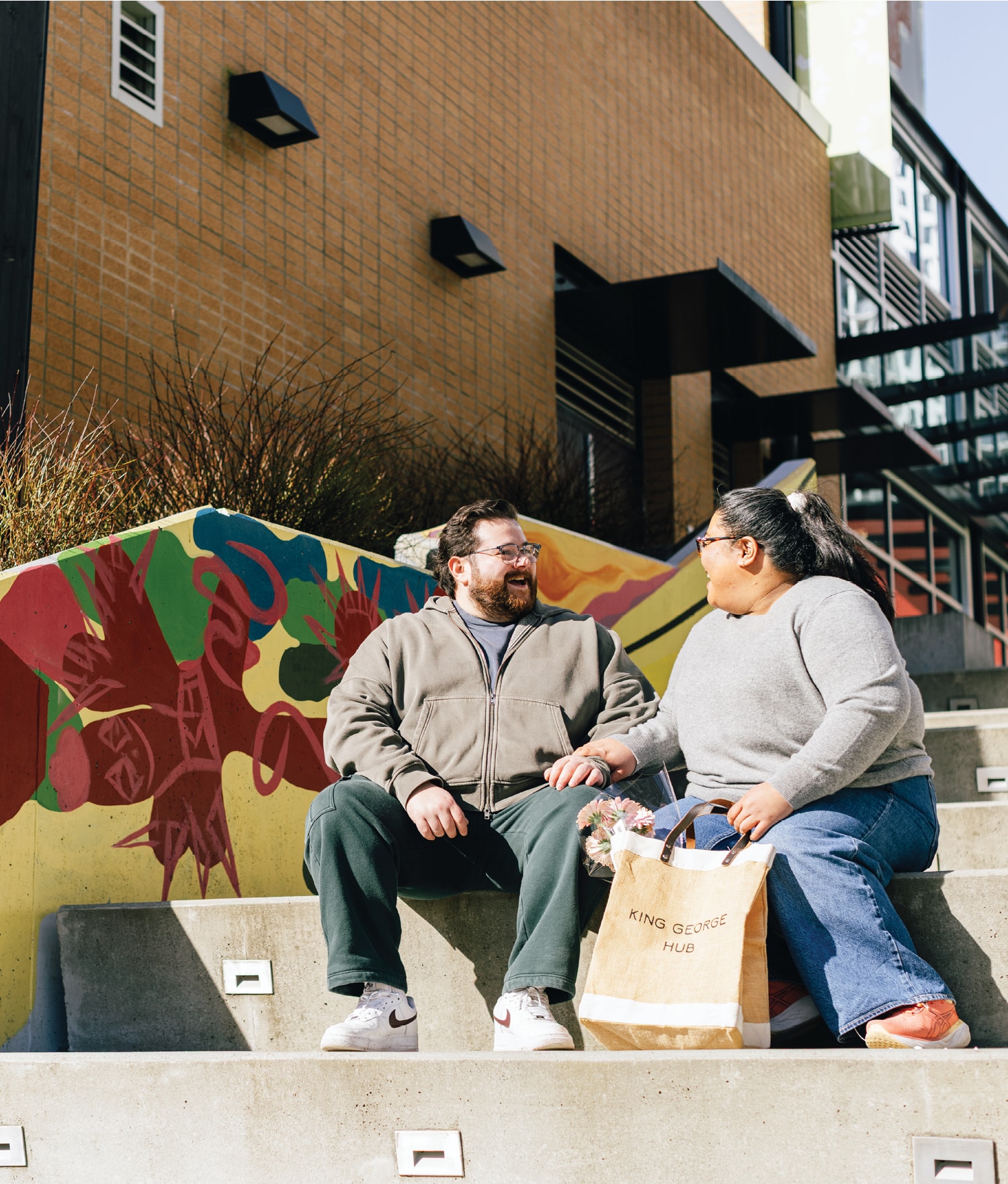 Two people sit on concrete steps beside a colorful graffiti wall and a bag reading “KING GEORGE HUB.”
